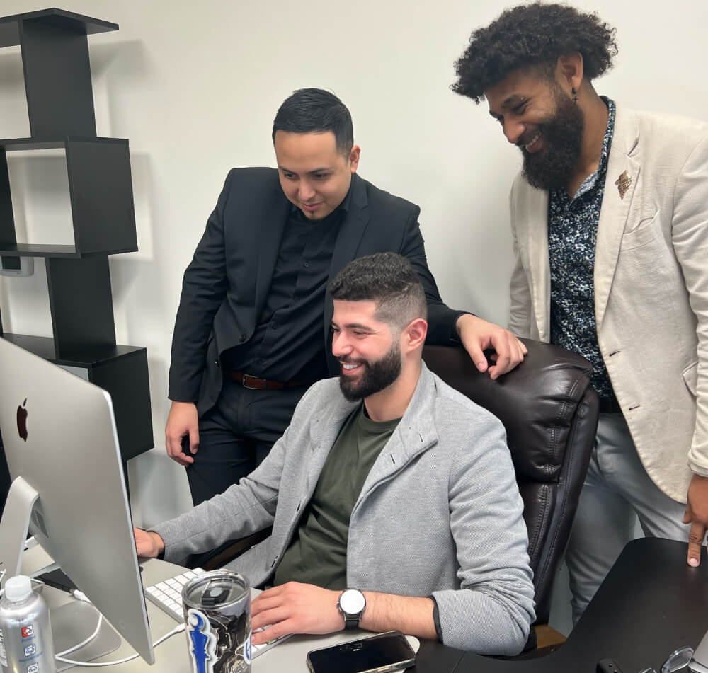 Three professional specialists smiling while reviewing a marketing campaign on a desktop computer in a modern office.